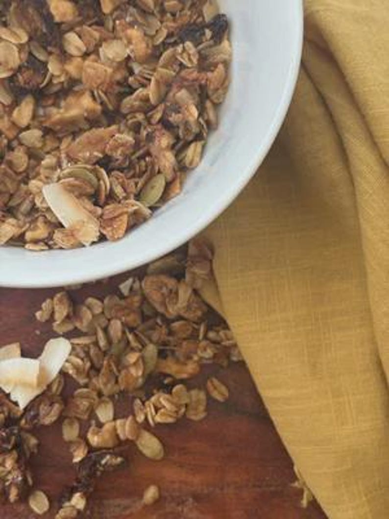 K&ouml;nnte beinhalten: Eine wei&szlig;e Sch&uuml;ssel gef&uuml;llt mit Granola aus ger&ouml;steten Haferflocken, Kokosflocken, K&uuml;rbiskernen und Trockenfr&uuml;chten. Loses Granola ist auf der dunklen Holzoberfl&auml;che neben der Sch&uuml;ssel verstreut, begleitet von einem gefalteten senfgelben Leinentuch.