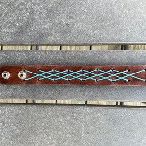 Op de afbeelding: Een bruine leren armband met turquoise kruissteken. De armband heeft zilveren drukkers aan elk uiteinde en is een stijlvol accessoire.