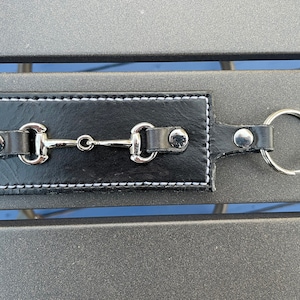 May include: Black leather keyring featuring a silver-coloured horse bit design. The keyring has white stitching around the edges and a silver key ring. The horse bit is a classic equestrian detail.