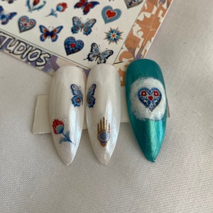 Può includere: Tre punte di unghie bianche e turchesi con adesivi per nail art colorati. Gli adesivi raffigurano una farfalla, una mano con un occhio e un cuore con un motivo floreale. Il foglio di adesivi mostra una varietà di disegni, tra cui farfalle, cuori e mani con occhi. Il foglio è etichettato "Anatolian Elegance Art Studios".