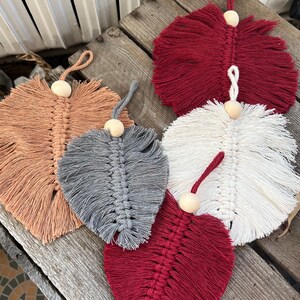 May include: Five macrame feather-shaped coasters in shades of red, white, gray, and peach. Each coaster features a central woven spine and a wooden bead for hanging. The coasters are displayed on a weathered wooden surface.