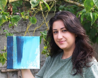 Puede incluir: Una mujer sostiene una pequeña pintura abstracta con pintura azul, verde y dorada. La pintura tiene una superficie texturizada.