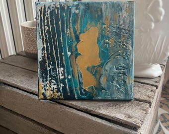 May include: Square abstract art piece with a textured surface. The artwork features a combination of teal, gold, and white paint. The painting is displayed on a wooden crate, with a white ceramic pot in the background.