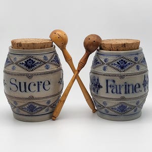 Peut inclure: Deux pots en céramique avec couvercles en liège et cuillères en bois. Les pots sont bleu clair avec des motifs floraux bleu foncé et les mots "Sucre" et "Farine". Les cuillères en bois sont croisées devant.