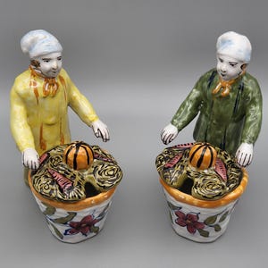 Peut inclure: Deux figurines en céramique de personnes portant des chapeaux et tenant des paniers remplis de légumes. Les paniers ont des couvercles avec un seul fruit orange sur le dessus. Les figurines sont peintes dans des couleurs vives, notamment le jaune, le vert, l'orange et le rouge.