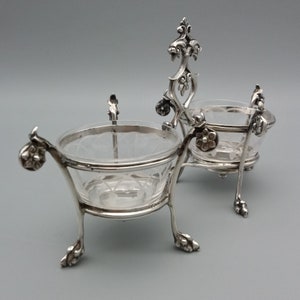 May include: A silver salt cellar set with two glass bowls on a decorative stand. The stand has a floral design and the bowls have a cut glass pattern.