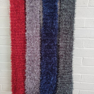 May include: Four fuzzy scarves in red, gray, blue, and dark gray hanging on a white brick wall.