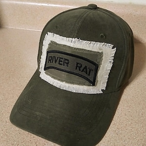 May include: Olive green baseball cap with a frayed patch that reads "RIVER RAT" in black lettering. The cap has a curved brim and a button on top. The patch is rectangular with a white border.
