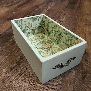 May include: A rectangular, light blue wooden box with a floral patterned interior and a decorative metal handle. The interior features a green background with pink and yellow floral accents. The box is set on a wooden surface.