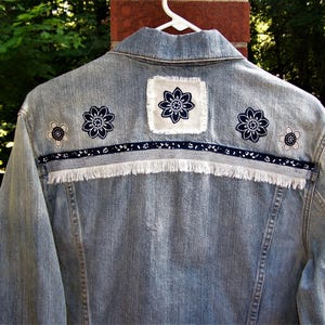 May include: Light blue denim jacket with floral embroidery. The back of the jacket features three navy blue and white flower designs, a white patch with a navy blue flower, and a navy blue ribbon with white fringe.