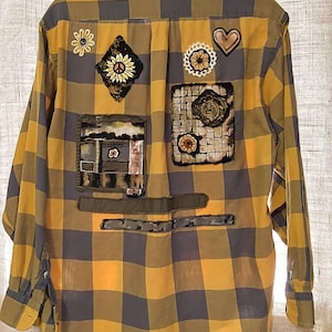 May include: A yellow and gray plaid flannel shirt with various fabric patches sewn on the back. The patches include a sunflower, a peace sign, a heart, and a floral design.