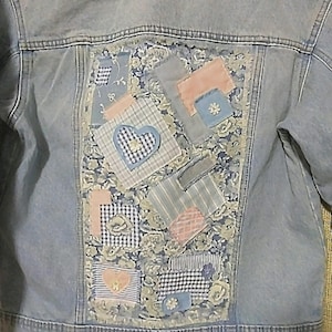May include: Light blue denim jacket with a patchwork design on the back. The patchwork features floral fabric, gingham, and solid patches in shades of blue, pink, and gray. Small flower embellishments are sewn onto some of the patches.