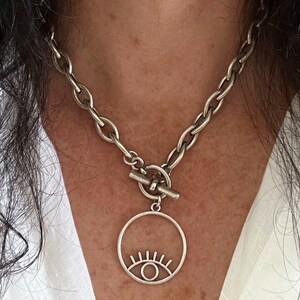 May include: A silver chain necklace with a toggle clasp and a circular pendant featuring a stylized eye design.