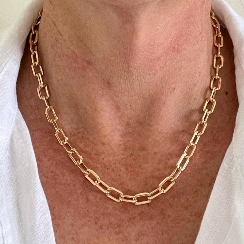 May include: A gold chain necklace with a square link design. The necklace is worn by a person with a white shirt.