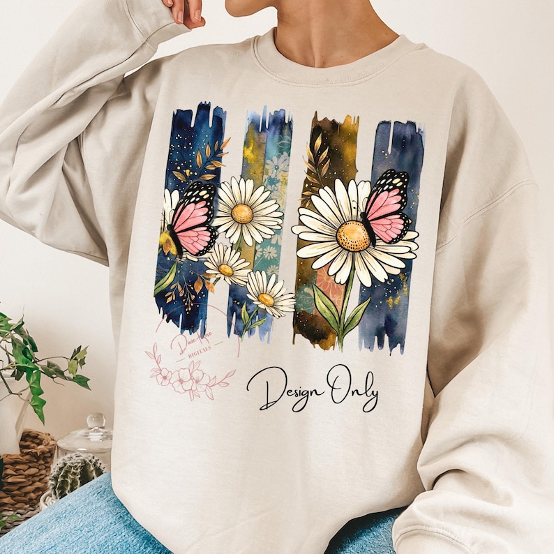 May include: Beige sweatshirt with a floral design. The design features white daisies, pink butterflies, and the text "Design Only" in black script. The background has vertical watercolor stripes in blue, yellow, and brown.