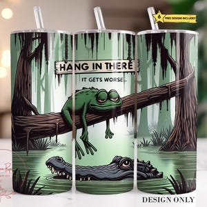 Puede incluir: Tres vasos de acero inoxidable con pajitas, con una escena de pantano. Una rana verde descansa sobre un tronco bajo un letrero que dice "HANG IN THERE, IT GETS WORSE". Un caimán nada en el agua. Los vasos tienen una combinación de colores verde y marrón.