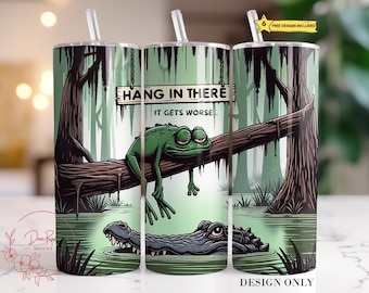 May include: A set of three stainless steel tumblers with straws, featuring a swamp scene. A green frog rests on a log under a sign that reads "HANG IN THERE, IT GETS WORSE." An alligator swims in the water below. The tumblers have a green and brown color scheme.