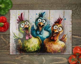 Peut inclure: Une planche à découper en verre colorée avec un design fantaisiste représentant trois poulets de dessin animé aux yeux écarquillés et au bec ouvert. Les poulets sont peints dans des tons de bleu, jaune et marron sur un fond de bois blanc.