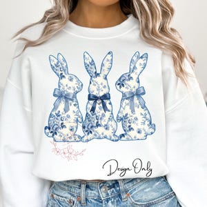 May include: White sweatshirt featuring three bunny silhouettes with blue floral patterns and bows. The bunnies have a vintage, porcelain-like aesthetic. The words "Design Only" are written in cursive below the bunnies.