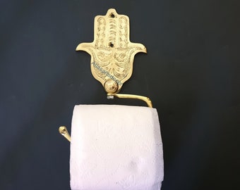 Puede incluir: Porta rollo de papel higiénico de metal dorado con un diseño decorativo de mano de Hamsa. El soporte está montado en la pared y tiene un rollo de papel higiénico blanco.