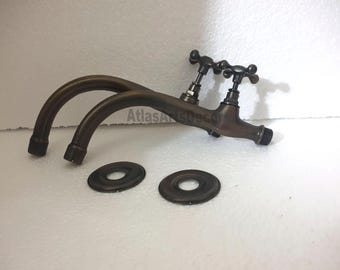 Puede incluir: Grifo doble de latón antiguo con dos caños curvos y dos placas decorativas redondas. El grifo tiene dos manijas negras con pomos en forma de cruz.