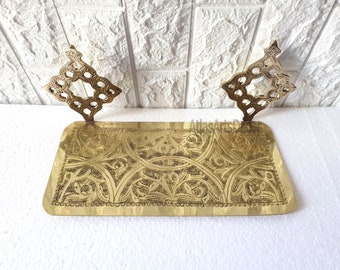 May include: A gold-colored metal tray with intricate floral designs etched into the surface. The tray has two decorative, ornate metal handles on either side.
