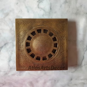 May include: A square brass drain cover with a circular design. The cover has a hammered texture and a decorative pattern around the center.