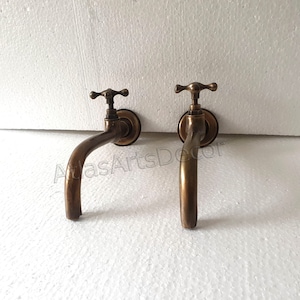 Puede incluir: Dos grifos de pared de latón antiguo con manijas en forma de cruz. Los grifos tienen un caño curvo y están montados sobre un fondo blanco.