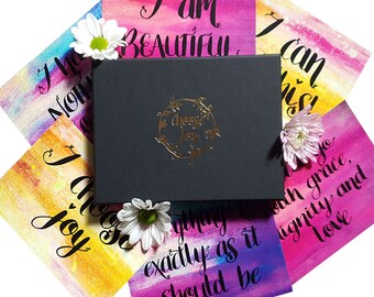 Puede incluir: Una caja negra con un logotipo en lámina dorada que dice "choose joy" rodeada de tarjetas coloridas con mensajes inspiradores escritos en caligrafía negra. Las tarjetas tienen fondos de acuarela en tonos de rosa, morado, amarillo y azul. Los mensajes en las tarjetas incluyen "Soy hermosa", "Elijo la alegría", "Puedo hacerlo", y "Todo está exactamente como debe ser".