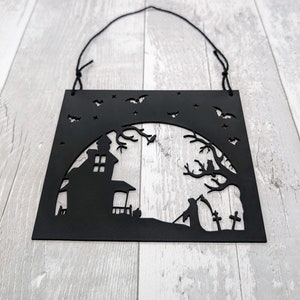 May include: Black Halloween wall decoration with a cut-out design of a haunted house, grim reaper, bats, and stars. The square sign has a string for hanging. The artwork is made of a solid black material.