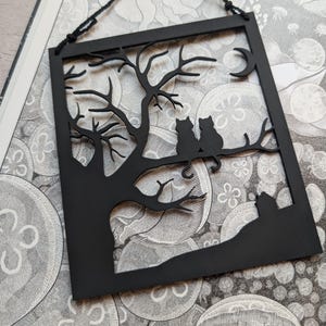 May include: A black, laser-cut paper artwork depicting a tree with two cats on a branch, a crescent moon, and a landscape silhouette. The framed piece is suspended by a black cord, ideal for wall decoration, and is made of paper.