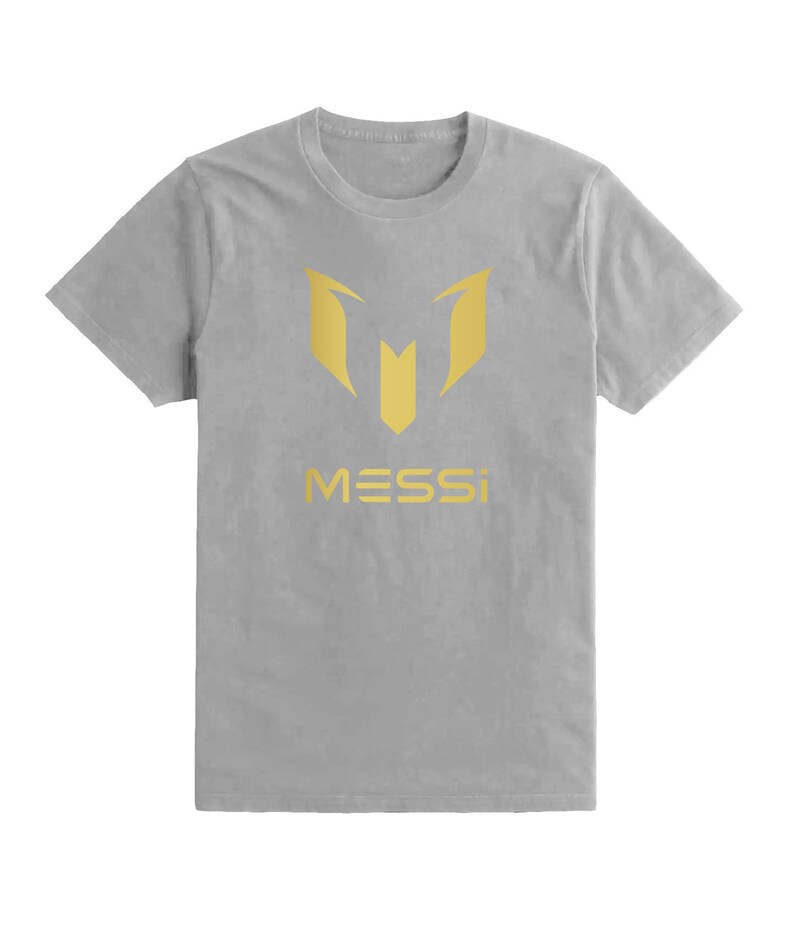 Op de afbeelding: Een grijs T-shirt met een gouden "M" logo en het woord "MESSI" gedrukt op de voorkant.