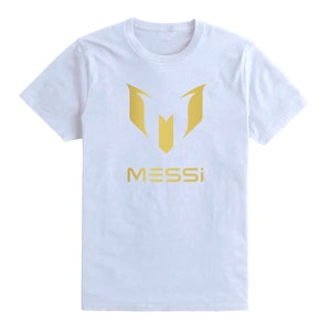 Op de afbeelding: Wit T-shirt met een gouden afbeelding van de letter "M" en het woord "MESSI" eronder.