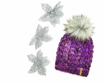May include: Purple and pink knitted beanie with a fluffy white pom pom. The beanie has a cable knit pattern and a brown leather tag with the word "Knit" on it. Three silver glitter poinsettia flowers are in the background.