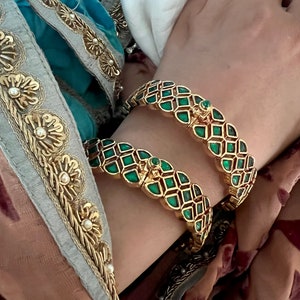 May include: Two gold bangles with green gemstone accents. The bangles are intricately designed with a repeating pattern of squares and diamonds.