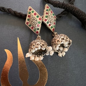 May include: Silver-toned earrings with a diamond-shaped top adorned with red and green gemstones. The earrings feature a bell-shaped bottom with dangling faux pearls. A black cord and a gold-colored cutout are also visible.