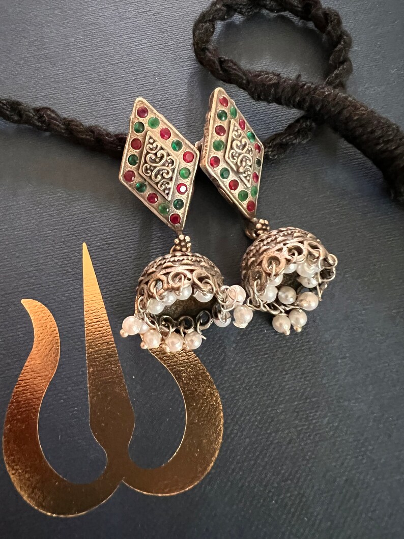 May include: Silver-toned earrings with a diamond-shaped top adorned with red and green gemstones. The earrings feature a bell-shaped bottom with dangling faux pearls. A black cord and a gold-colored cutout are also visible.