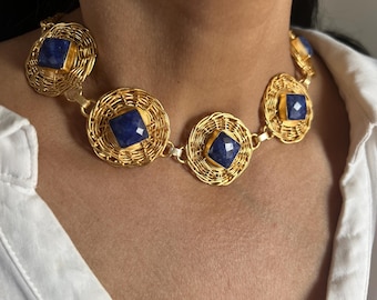 May include: A gold-toned necklace featuring square, deep blue gemstones set within woven gold circular frames. The necklace is composed of multiple linked segments, creating a statement piece. The necklace is worn over a white shirt.