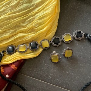 May include: A silver necklace with black and yellow square stones and matching earrings. The necklace is on a yellow fabric background.