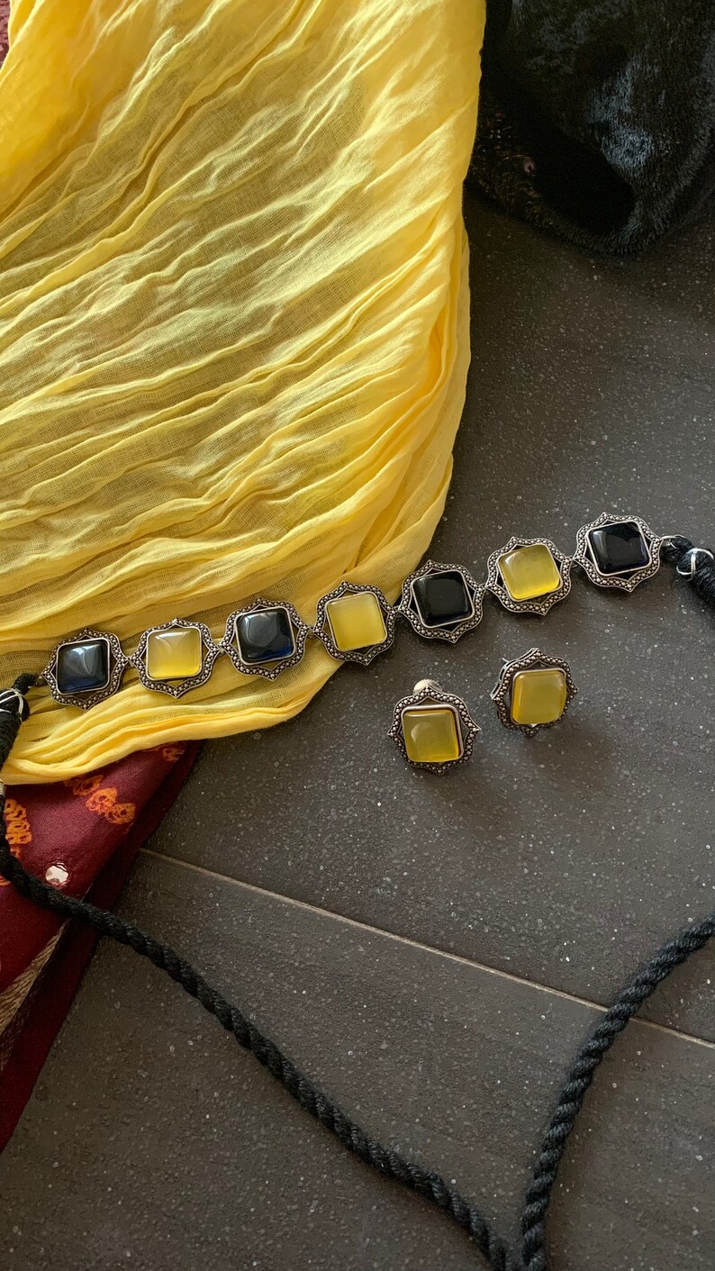 May include: A silver necklace with black and yellow square stones and matching earrings. The necklace is on a yellow fabric background.
