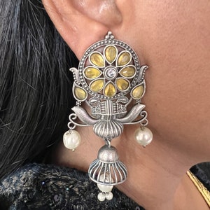 May include: Silver earrings with a floral design featuring yellow and white stones. The earrings have a dangling bell charm and pearl accents.