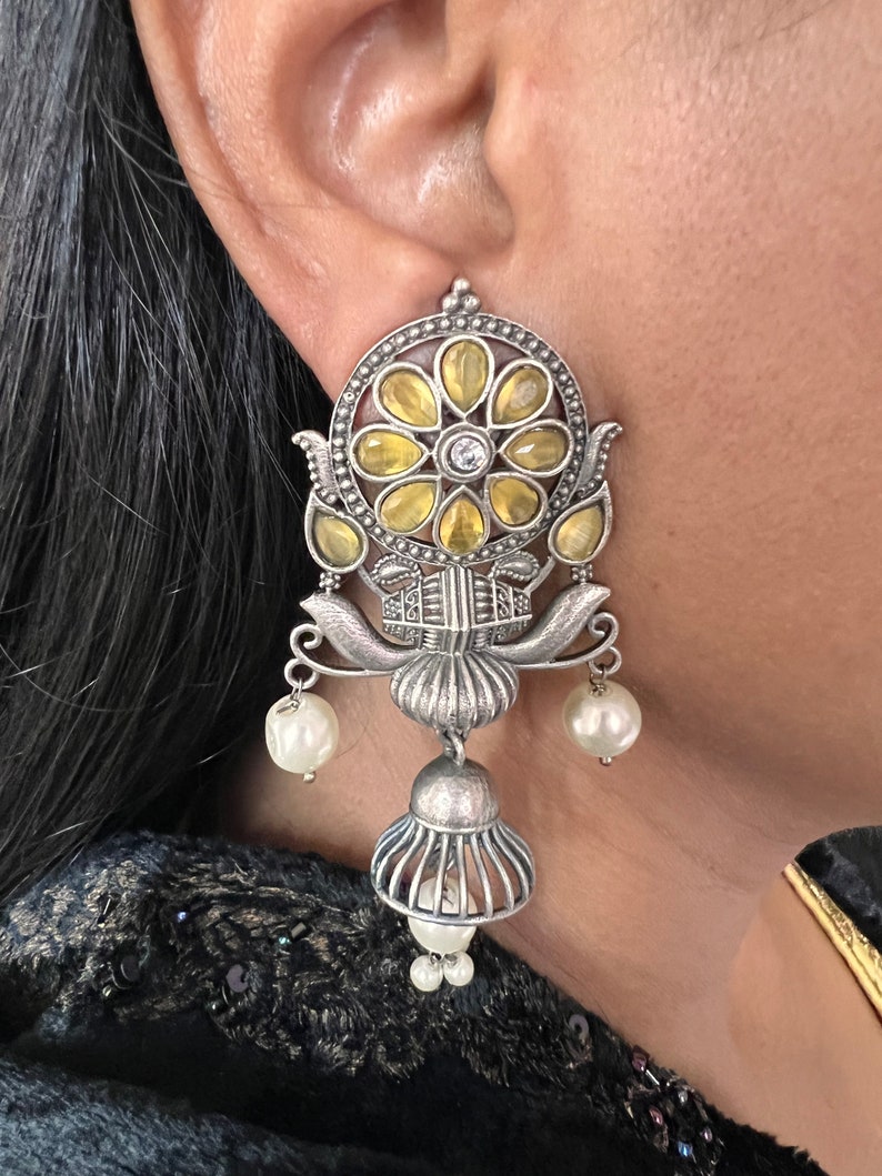 May include: Silver earrings with a floral design featuring yellow and white stones. The earrings have a dangling bell charm and pearl accents.