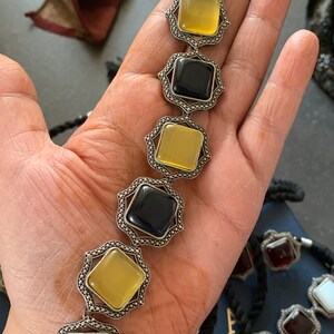 May include: A silver necklace with alternating black, yellow, and blue square gemstones. The necklace is made of a silver chain with a black cord.