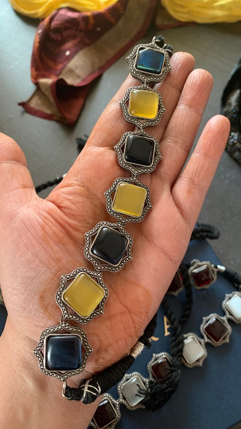 May include: A silver necklace with alternating black, yellow, and blue square gemstones. The necklace is made of a silver chain with a black cord.