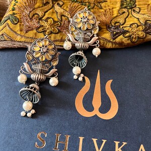 May include: A pair of silver earrings with a floral design and pearl accents. The earrings are on a blue background with the word "SHIVKA" in gold lettering.