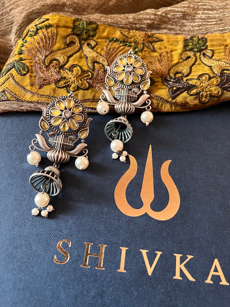 May include: A pair of silver earrings with a floral design and pearl accents. The earrings are on a blue background with the word "SHIVKA" in gold lettering.