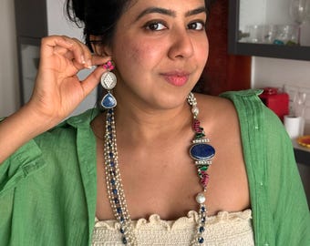 May include: A woman wearing a long, layered necklace with blue and white beads, and a matching earring. The earring has a pink stud, a silver disc, and a blue teardrop. The necklace has a large blue oval stone.