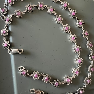 Peut inclure: Un collier en chaîne argent avec des breloques en forme de fleurs roses. Le collier a deux fermoirs et est fait de maillons délicats.