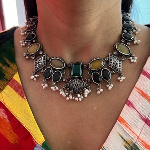 May include: A silver-toned necklace with a variety of gemstones in oval, square, and diamond shapes. The necklace features black, yellow, green, and red stones, with small white pearl accents. The necklace is worn against a colorful patterned top.