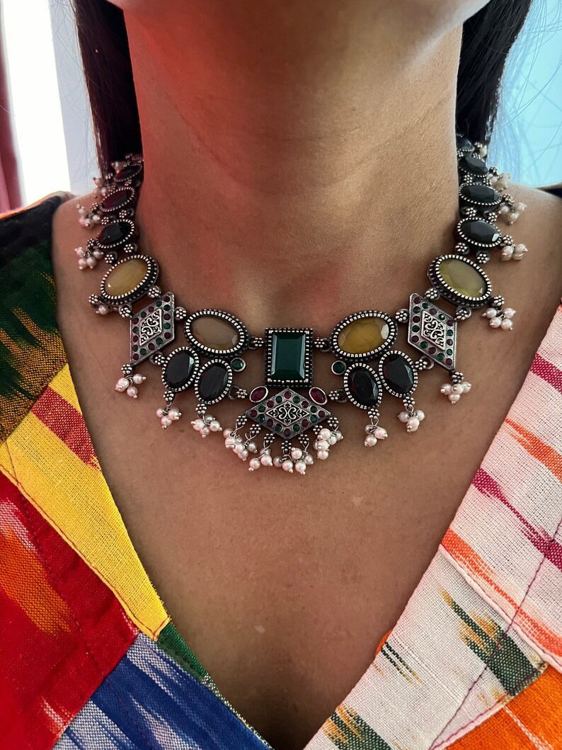 May include: A silver-toned necklace with a variety of gemstones in oval, square, and diamond shapes. The necklace features black, yellow, green, and red stones, with small white pearl accents. The necklace is worn against a colorful patterned top.